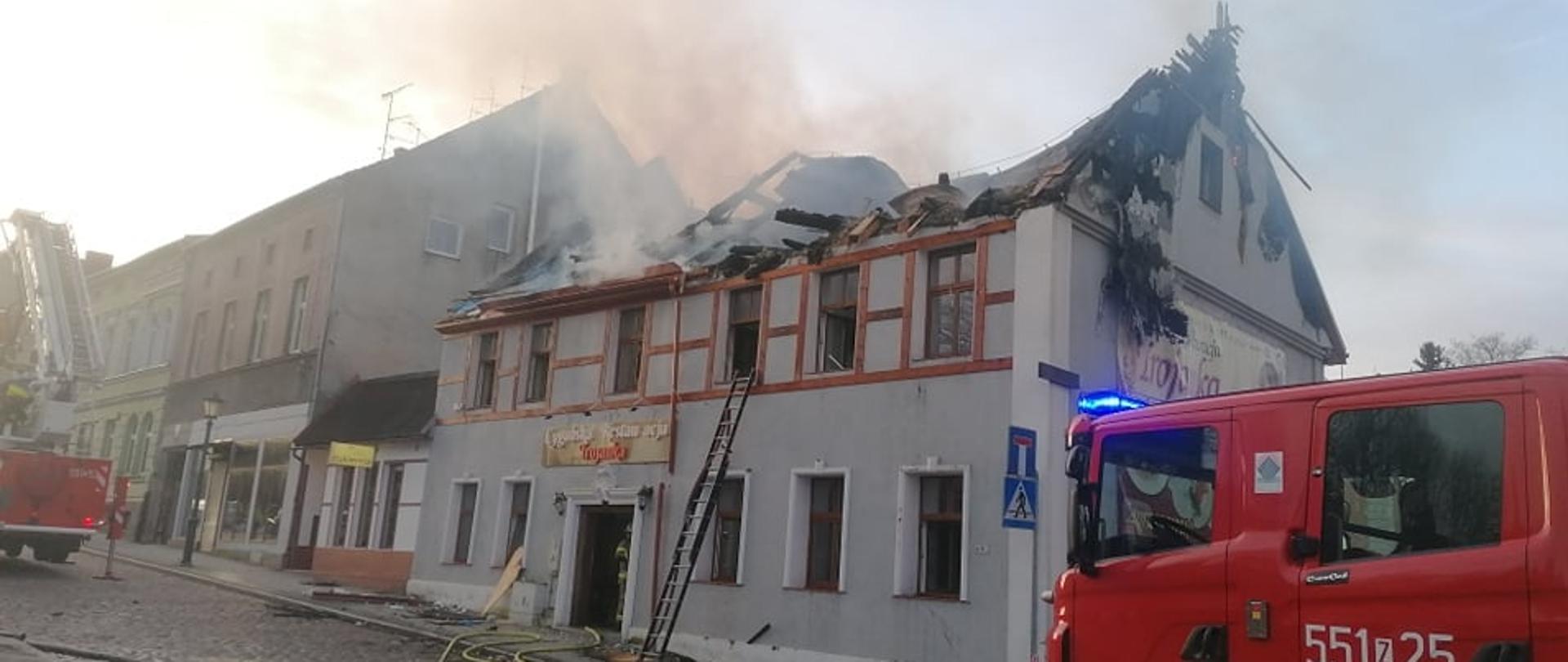 Zdjęcie przedstawia budynek restauracji dwukondygnacyjne podczas pożaru poddasza. Budynek koloru białego znajduje się centralnie w środku zdjęcia do budynku wchodzą linie gaśnicze obok budynku znajdują się dwa wozy strażackie. 