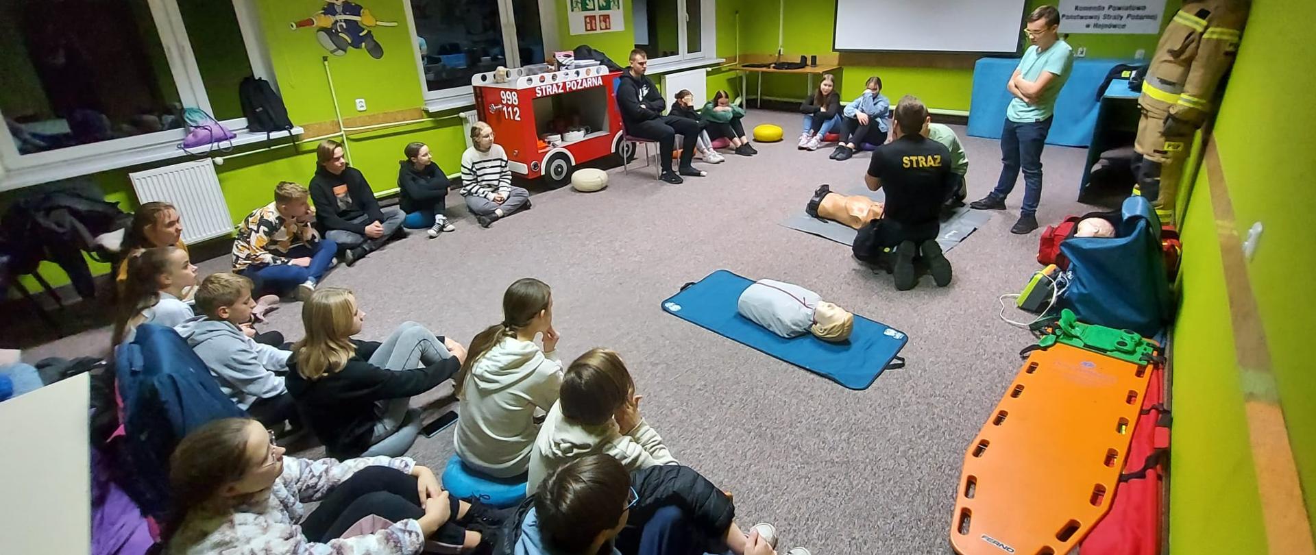 W sali z zielonymi ścianami w koło siedzi kilkanaście dzieci po turecku. Po środku na podłodze leżą dwa manekiny. Przy jednym manekinie z jednym dzieckiem ćwiczy strażak w czarnym mundurze.