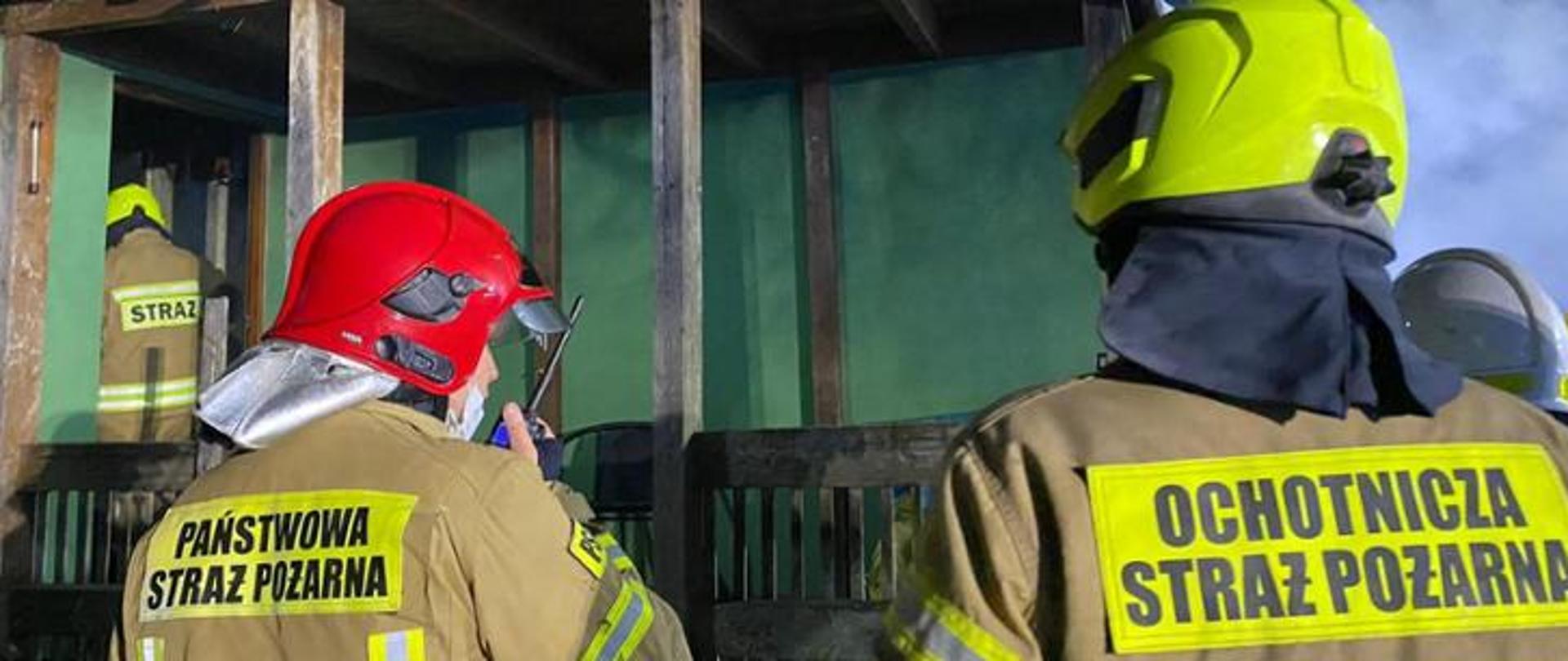 Na pierwszym planie dwaj strażacy w ubraniach specjalnych. Strażacy odwróceni do aparatu tyłem. Strażak po prawej stronie na plecach ma napis Państwowa Straż pożarna, a po lewej stronie zdjęcia strażak z napisem na plecach Ochotnicza Straż Pożarna