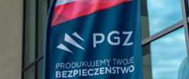 Zbliżenie na powiewającą na zewnątrz flagę Polskiej Grupy Zbrojeniowej (PGZ) w barwach ciemnoniebieskiej, białej i czerwonej, z napisem "PGZ PRODUKUJEMY TWOJE BEZPIECZEŃSTWO". W tle widać przeszklony budynek.