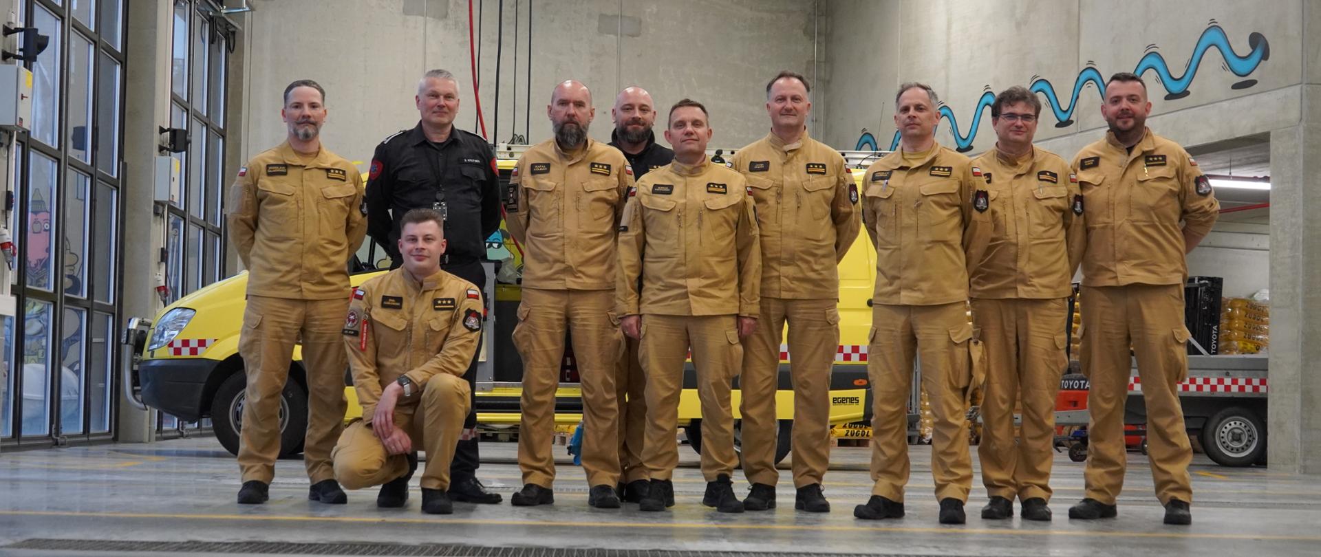 Grupa strażaków PSP pozuje do wspólnego zdjęcia w garażu jednostki ratowniczej. W tle widoczny jest żółty pojazd ratowniczy oraz wyposażenie techniczne.