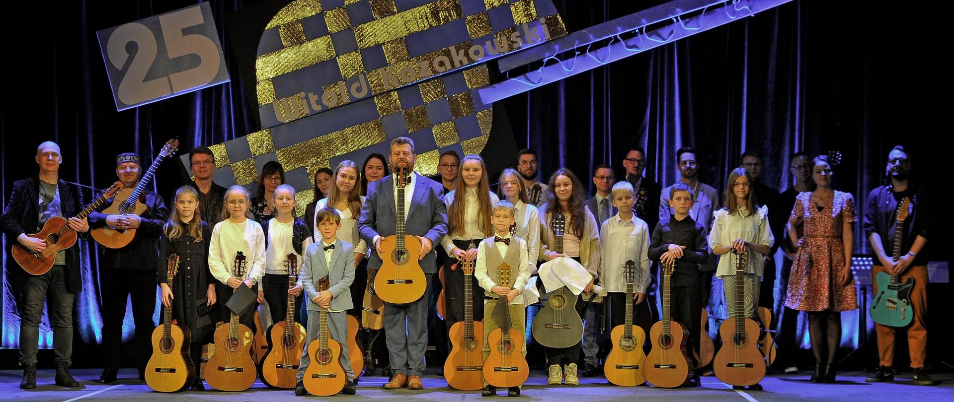 Zdjęcie grupowe wykonane na scenie w Kłodzkim Ośrodku Kultury, wszystkie osoby na zdjęciu trzymają gitarę, a w tle widoczna jest grafika gitary z napisem 25 Witold Kozakowski