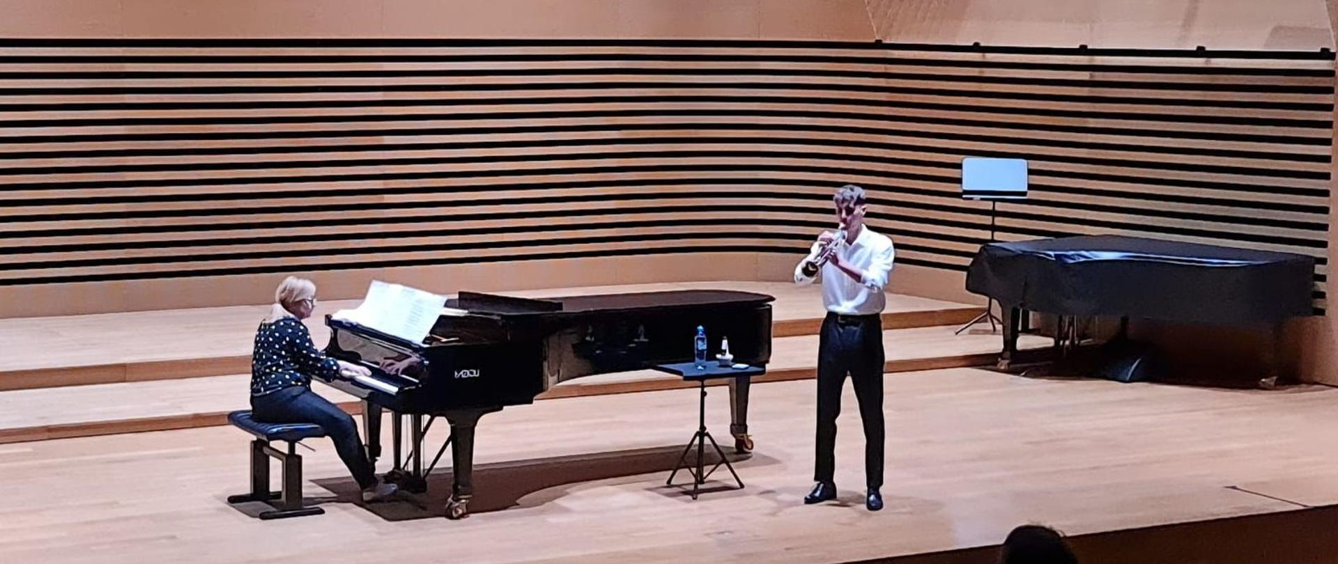 Chłopak gra na trąbce, kobieta akompaniuje na fortepianie.