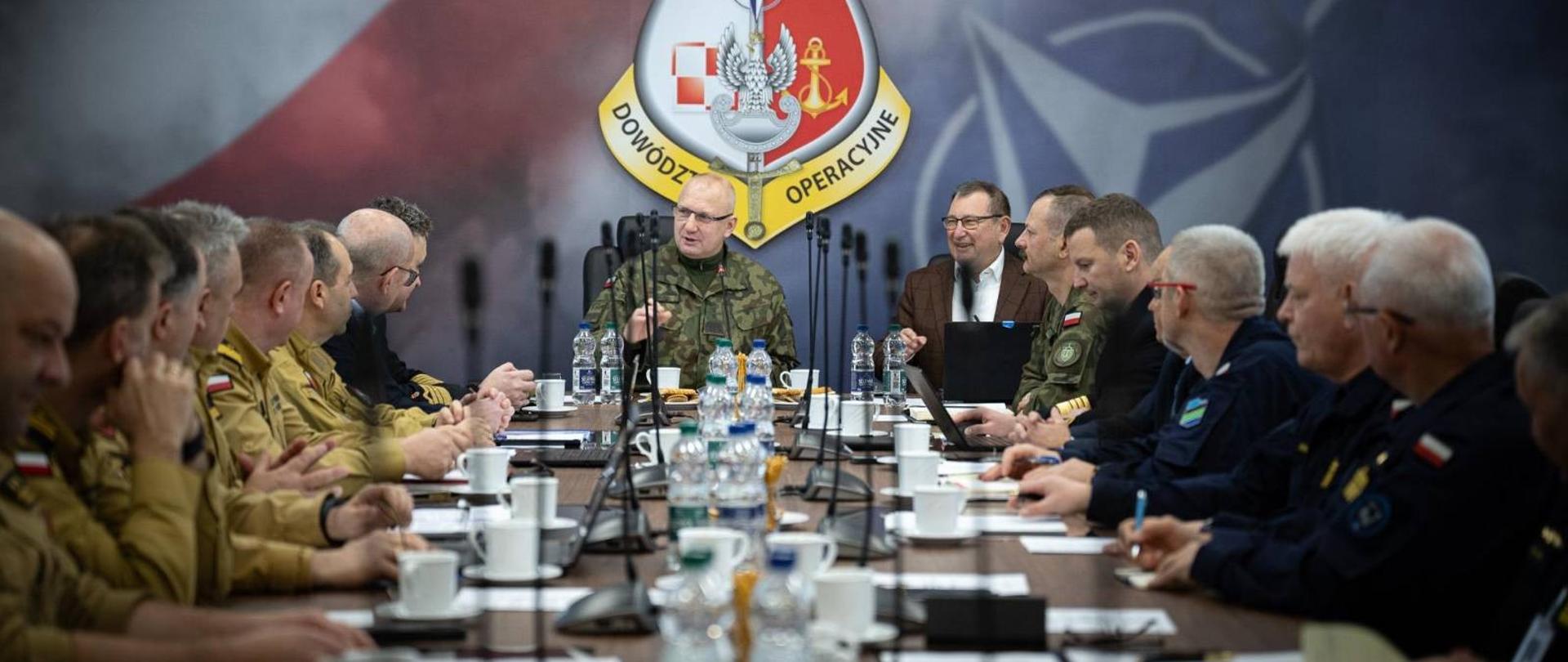 Długi stół przy którym po obu stronach siędzą funkcjonariusze. na stole filiżanki i laptopy.