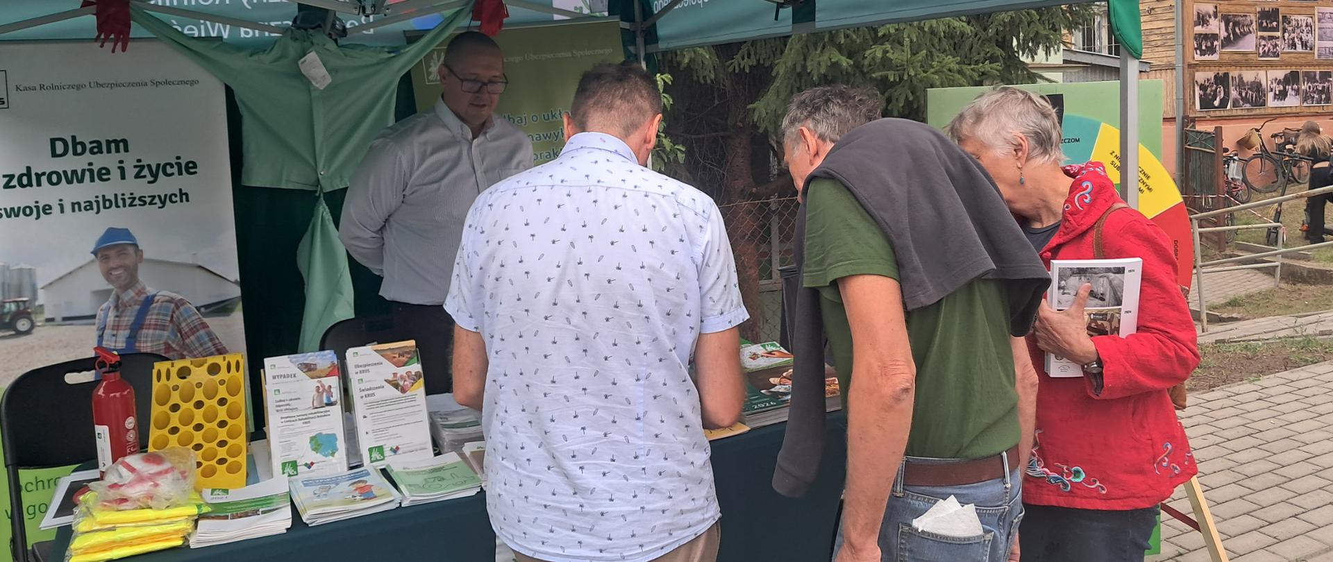 Pracownik oddziału stoi pod namiotem, w którym zorganizowano stoisko informacyjno-prewencyjne dla uczestników Dożynek. Przed namiotem od strony prawej ustawiono koło bezpieczeństwa. W namiocie po lewej stronie stoi stolik z ulotkami oraz materiałami prewencyjnymi, po prawej ustawiono roll-up i ekspozycję ochronnej odzieży osobistej. Pracownik stojący przed stoiskiem udziela informacji mężczyźnie i kobiecie odwiedzającym stoisko.