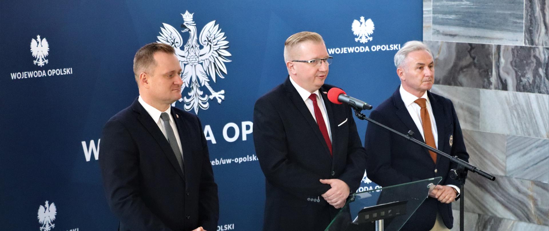 Trzech mężczyzn stojących przed mikrofonem na niebieskim tle 