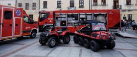 Dwa czerwone pojazdy ratownicze – quad i pojazd wielokołowy (typu amfibia) – stoją na brukowanym rynku. W tle zaparkowane są duże wozy strażackie i ambulanse na tle zabytkowych kamienic.