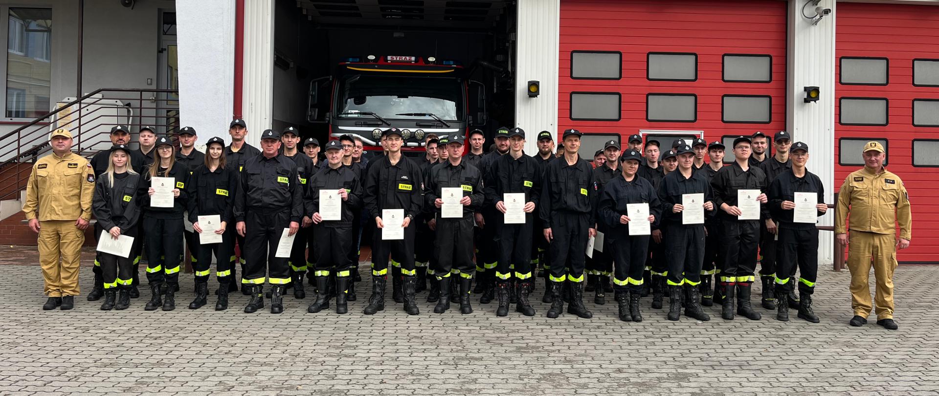Plac przed garażami Jednostki Ratowniczo Gaśniczej we Włoszczowie, na tle garaży w trójszeregu stoją słuchacze wraz z dowództwem JRG, którzy właśnie odebrali zaświadczenia ukończenia szkolenia podstawowego strażaka ratownika OSP.