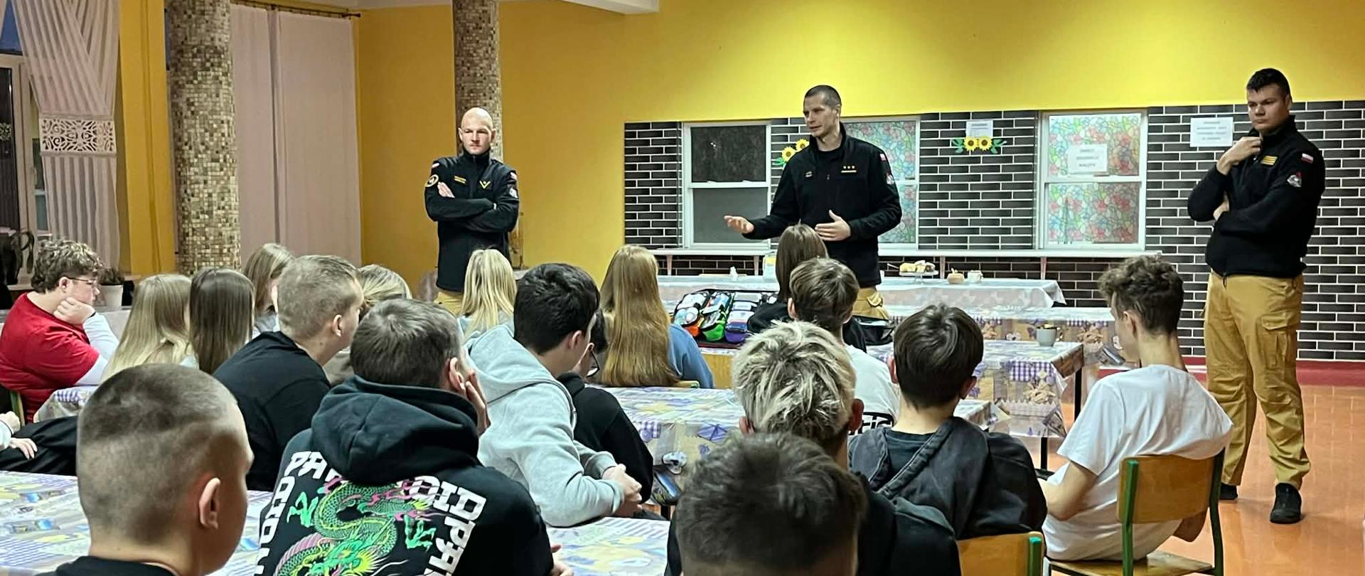 Na głównym planie jest trzech strażaków. Strażak będący pośrodku opowiada, gestykulując dłońmi. Uczniowie ukazani od pleców, zwróceni w kierunku prelegenta. 