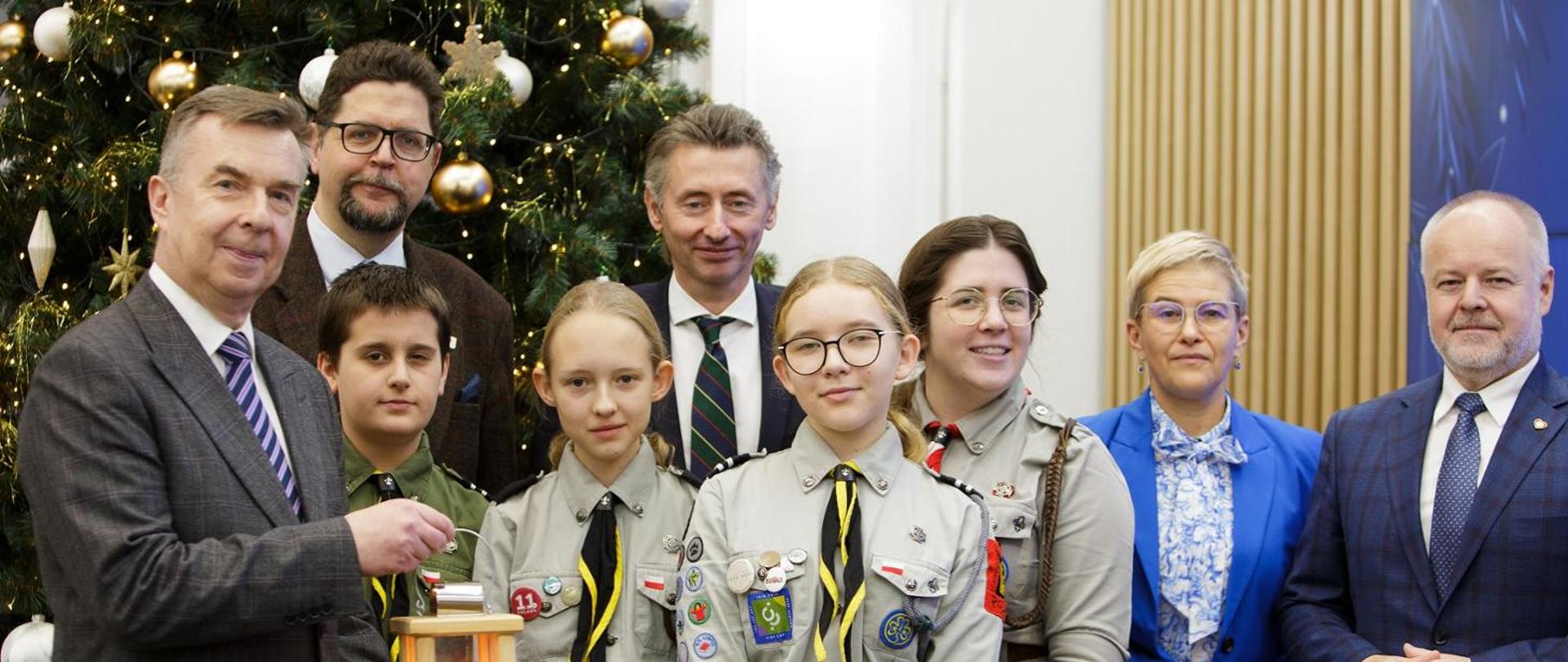 Pod ozdobioną bombkami i łańcuchami choinką stoi minister Wieczorek, wiceministrowie i czworo harcerzy w mundurach.