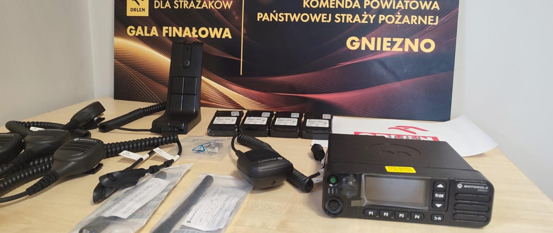 Czarny sprzęt łączności: radia, baterie, mikrogłośniki, anteny leżą na jasnym stole.