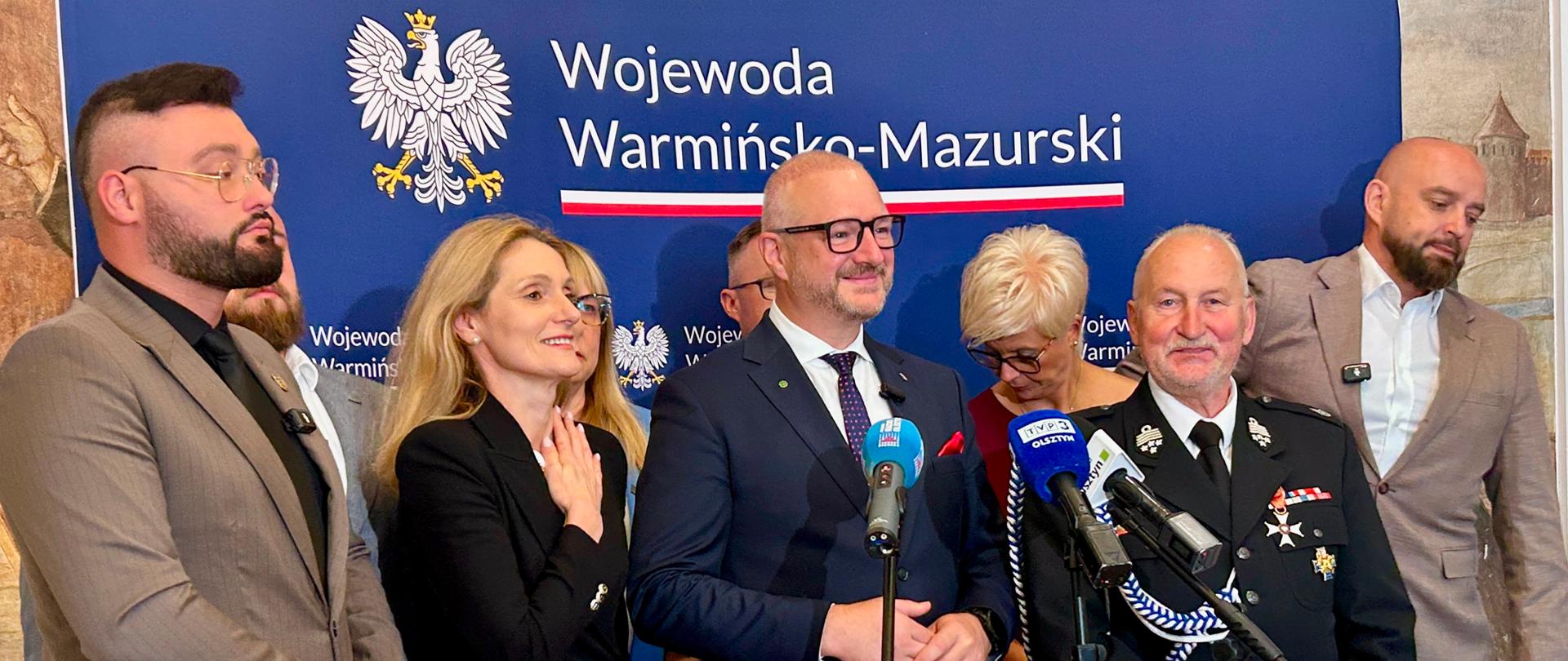 Ujęcie z boku przedstawiające grupę oficjeli oraz wojewodę warmińsko-mazurskiego Radosława Króla podczas briefingu prasowego. Uczestnicy stoją wspólnie na tle ścianki, uśmiechają się i pozują do zdjęcia.