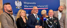 Ujęcie z boku przedstawiające grupę oficjeli oraz wojewodę warmińsko-mazurskiego Radosława Króla podczas briefingu prasowego. Uczestnicy stoją wspólnie na tle ścianki, uśmiechają się i pozują do zdjęcia.