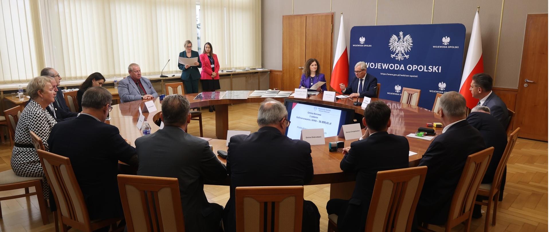 Zdjęcie grupowe. Przedstawicie gmin i Wojewoda Opolski zasiadają przy okrągłym stole w sali im. Braci Kowalczyków Opolskiego Urzędu Wojewódzkiego.