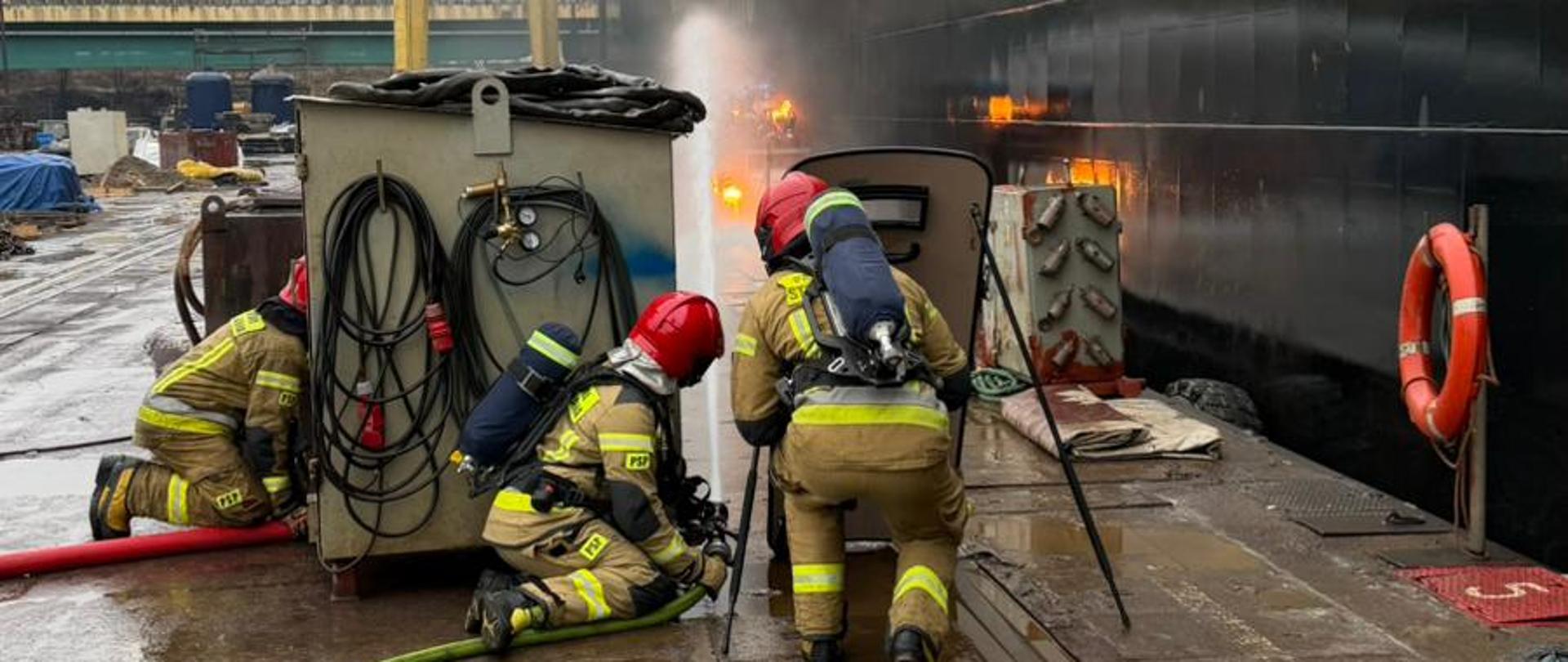 Fotografia przedstawia czterech strażaków w pełnym umundurowaniu bojowym (ubrania specjalne, hełmy, aparaty oddechowe) podczas akcji gaśniczej w porcie lub stoczni. Strażacy klęczą lub pochylają się na mokrym, betonowym nabrzeżu, operując sprzętem gaśniczym (wąż strażacki, prądownica). W tle po prawej stronie znajduje się duży, czarny kadłub statku, z którego unosi się dym lub para. Po lewej stronie widoczne są żółte żurawie portowe. Na nabrzeżu leżą węże strażackie i widać odbicia światła w kałużach wody.