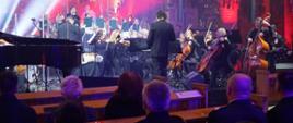 Orkiestra gra koncert na instrumentach w ławach siedzą zaproszeni goście i słuchają.