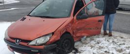 Na zdjęciu widać rozbity przód samochodu osobowego. Obok auta stoi odwrócona kobieta. W tle stoi strażak oraz samochód ratowniczo-gaśniczy. Samochód stoi na poboczu drogi krajowej