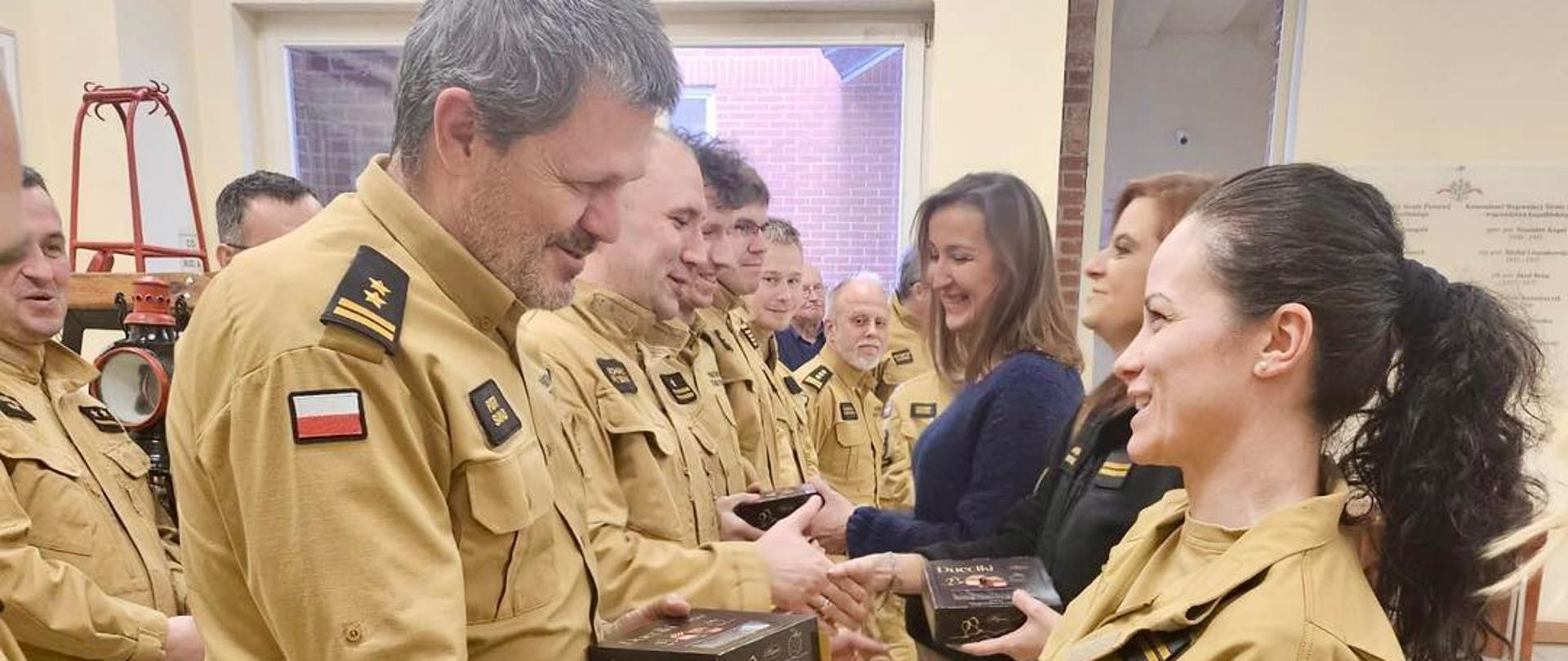 Strażacy w mundurach stoją w dwóch rzędach w pomieszczeniu podczas uroczystości. Każdy z nich otrzymuje pudełko z upominkiem. Na pierwszym planie mężczyzna w mundurze ściska dłoń kobiecie również ubranej w mundur strażacki.