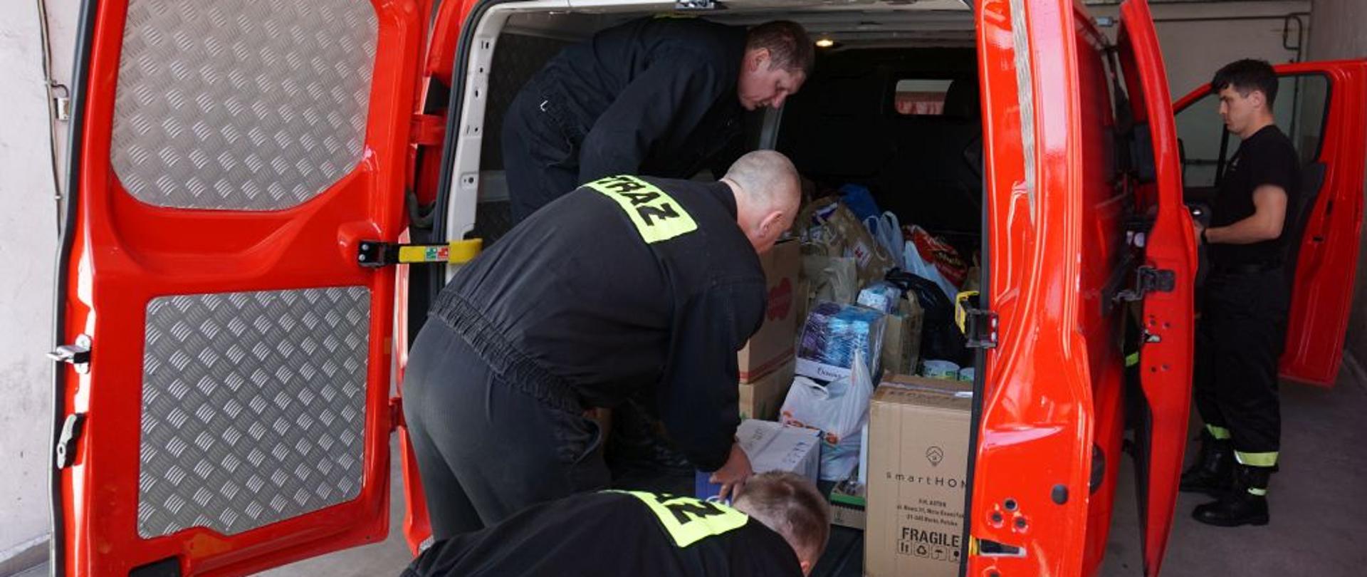 3 mężczyzn w czarnych mundurach strażackich pakuje do bagażnika czerwonego busa żywność i środki czystości.