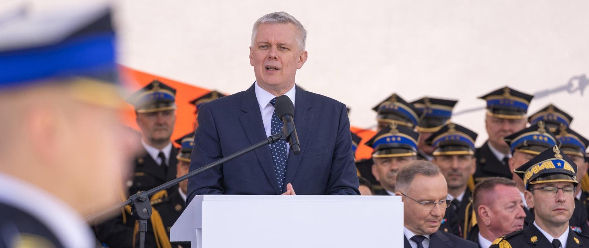 Minister Tomasz Siemoniak podczas Centralnych Obchodów Dnia Strażaka. Szef MSWiA stoi przed mównicą. W tle po prawej zsiada Prezydent RP i strażacy. Na pierwszym planie po lewej strażak.