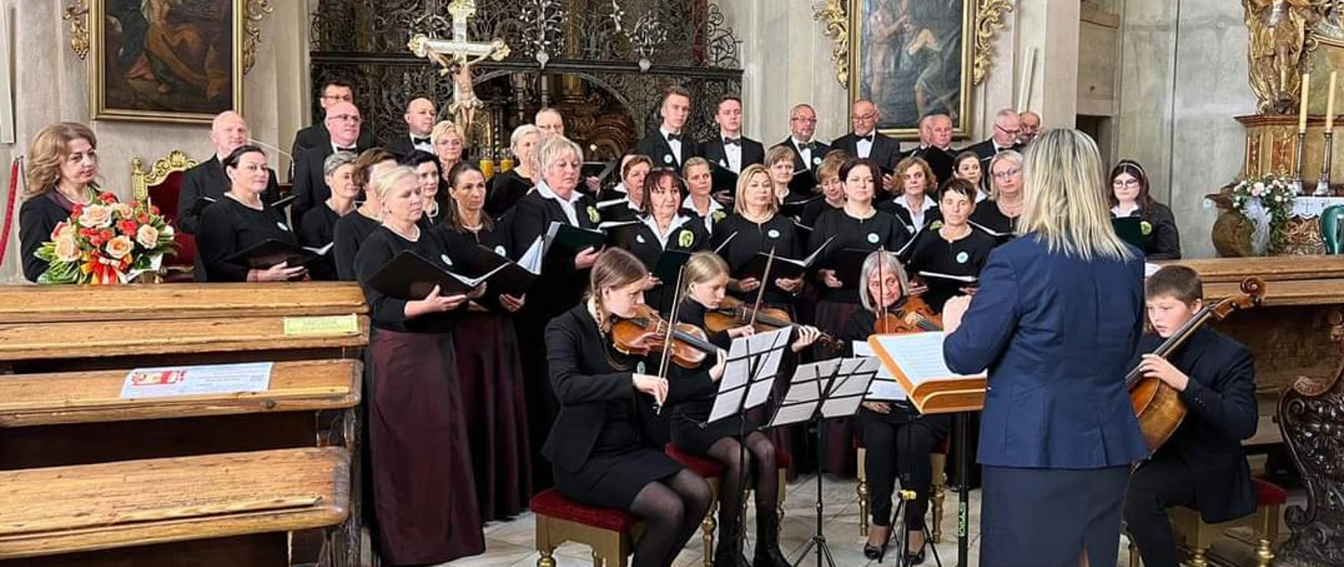 Grupa śpiewających osób, z osobami akompaniującymi na skrzypcach i wiolonczeli oraz osoba dyrygująca na pierwszym planie na tle ołtarza w kościele