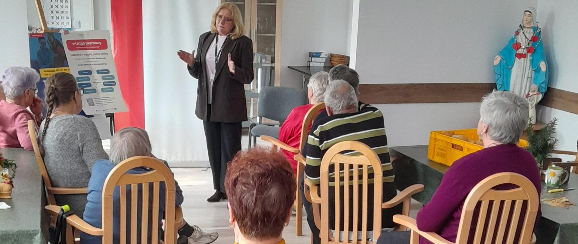 Osoby starsze siedzą na krzesłach. Przed nimi stoi pracownica urzędu skarbowego