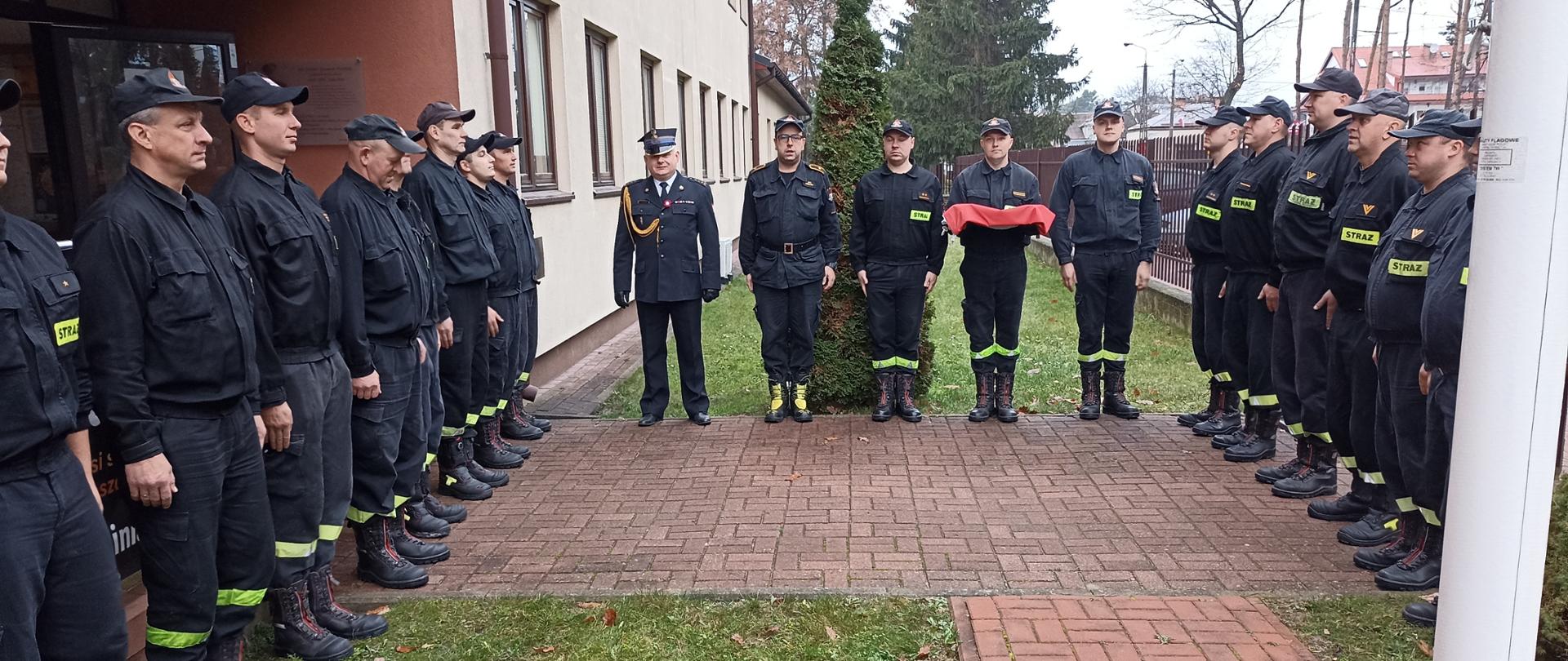 Zdjęcie przedstawia strażaków ustawionych w dwóch szeregach, zwróconych przodem do siebie. W centralnym punkcie fotografii znajduje się ubrany w mundur galowy Komendant Powiatowy PSP. W trakcie uroczystej zmiany służby strażacy wciągnęli flagę państwową na maszt oraz odśpiewali hymn Polski.