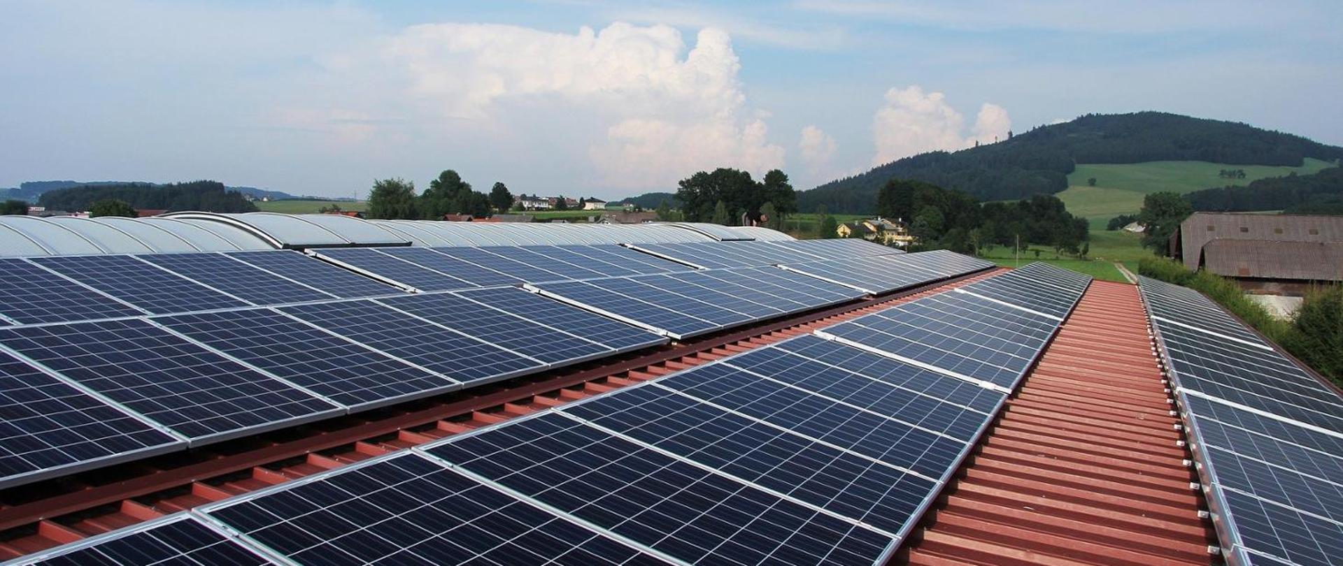 Szereg paneli fotowoltaicznych zamontowanych na czerwonym metalowym dachu z widokiem na zielone wzgórza i zabudowania w tle, pod częściowo zachmurzonym niebem.