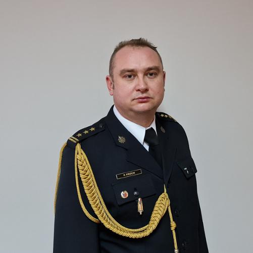 Na zdjęciu widać mężczyznę w mundurze galowym, stojącego na tle jasnej ściany. Ma on krótkie, jasne włosy i patrzy prosto przed siebie. Ubrany jest w ciemny mundur, ozdobiony złotymi pagonami z trzema gwiazdkami oraz złotym, plecionym sznurem przewieszonym przez lewe ramię. Na lewej piersi munduru widoczne są przypinki. Mężczyzna ma poważny wyraz twarzy.