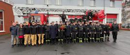 Zdjęcie przedstawia strażaków pozujących do zdjęcia. Strażacy stoją na placu przed drabiną mechaniczną oraz budynkiem komendy.