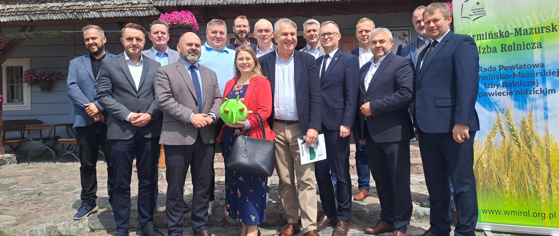 Grupa ludzi stoi pozując do zdjęcia. pośrodku grupy mężczyzn w garniturach stoi kobieta w czerwonej marynarce z zieloną maskotką w ręku. W tle dom kryty gontem, z prawej strony rollup WMIR