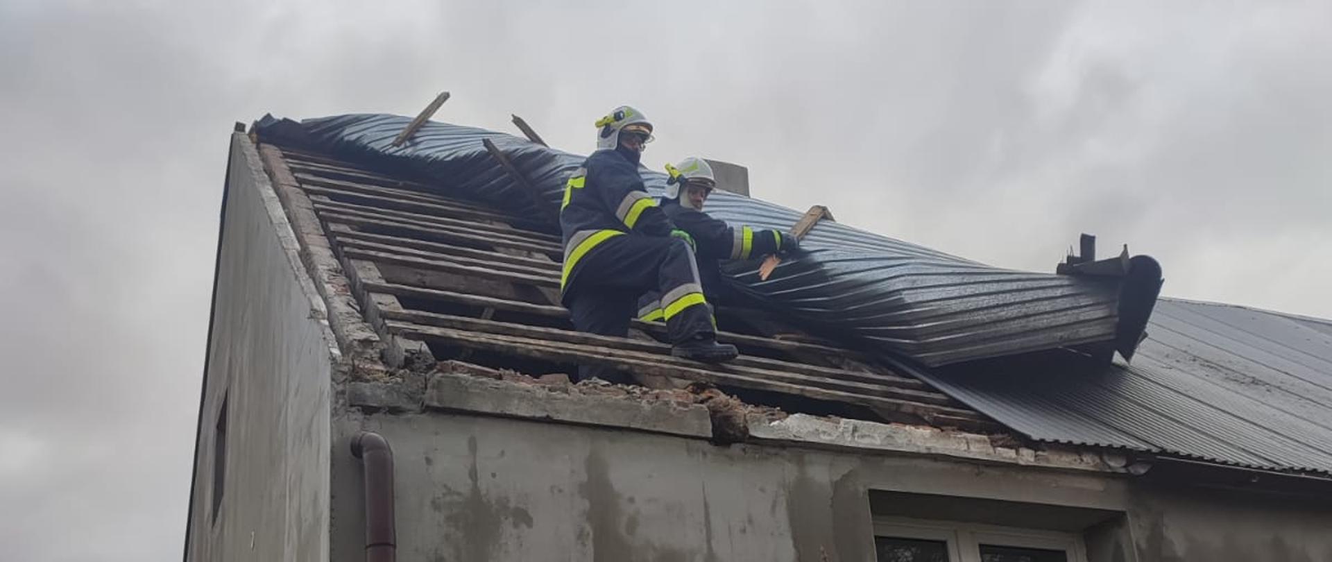 Dwóch strażaków w pełnym umundurowaniu naprawia częściowo zerwane poszycie na dachu domu jednorodzinnego. Pochmurna pogoda.