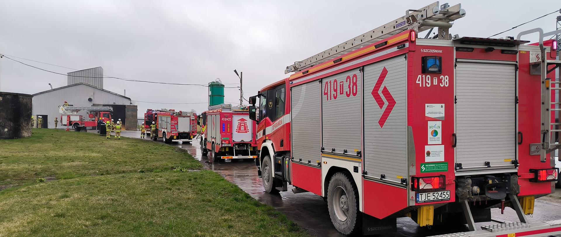 Zdjęcie przedstawia zaparkowane pojazdu pożarnicze przy działaniach gaśniczych. W tle budynek suszarni.