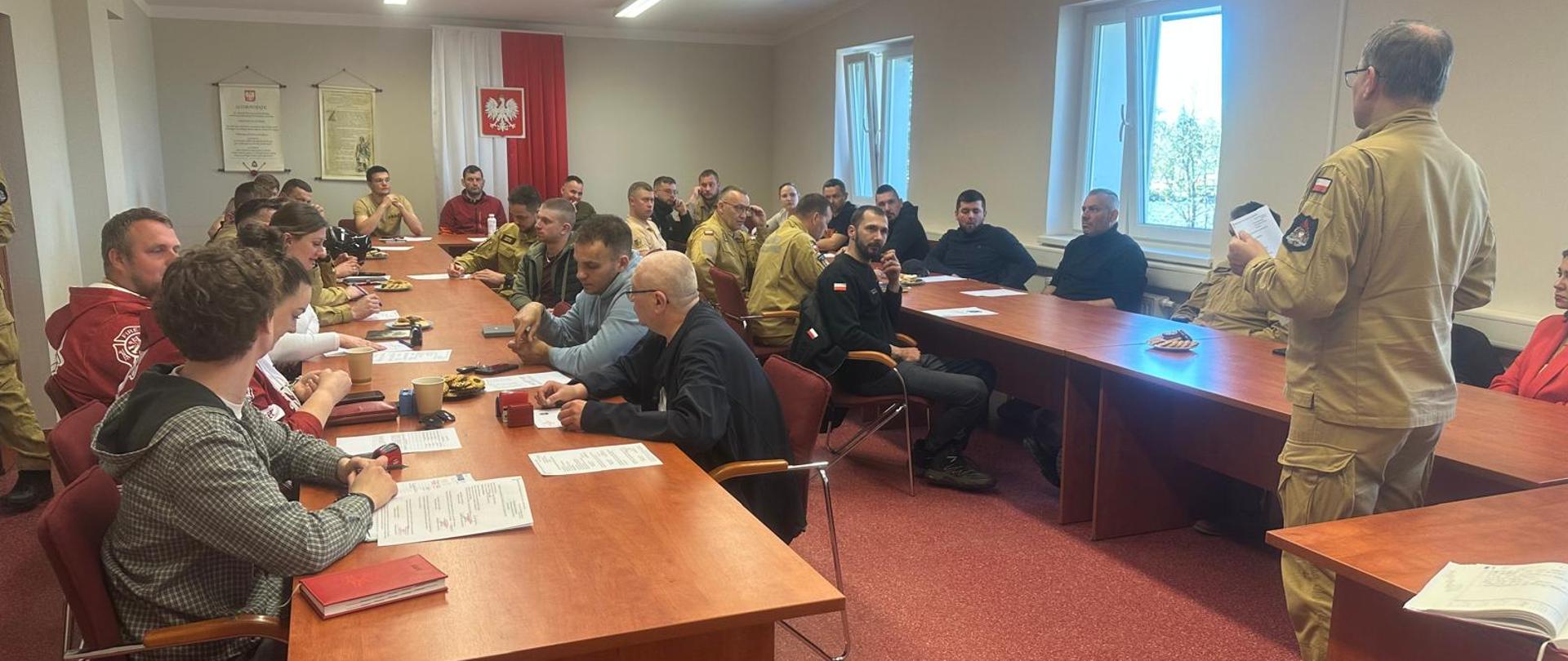 Uczestnicy spotkania strażackiego siedzą przy stołach w sali konferencyjnej, słuchając komendanta powiatowego, prowadzącego stojącego z przodu pomieszczenia. W spotkaniu bierze udział kilkadziesiąt osób, członków Zarządu jednostek OSP, część z nich jest w mundurach strażackich, część w ubraniach cywilnych. Na stołach leżą dokumenty, notesy i kubki. W tle widoczna jest biało-czerwona flaga z godłem Polski oraz okna doświetlające salę.