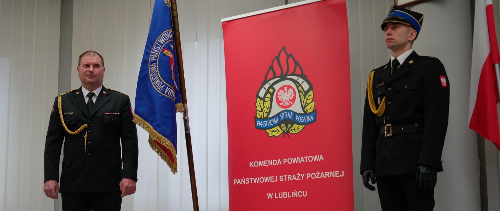 Zdjęcie zrobione w pomieszczeniu strażnicy na tle na tle godła Rzeczpospolitej Polskiej, Sztandaru Komendy Powiatowej Państwowej Straży Pożarnej w Lublińcu, dwóch Flag Rzeczpospolitej Polskiej oraz baneru Komendy Powiatowej PSP w Lublińcu. Na pierwszym planie, po lewej stronie funkcjonariusz odchodzący na zaopatrzenie emerytalne ubrany w umundurowanie galowe.