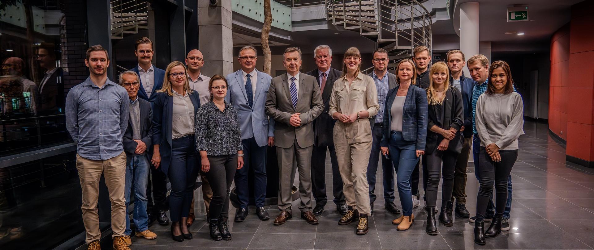 Zdjęcie zbiorowe, duża grupa ludzi stoi na wyłożonym szarymi kamiennymi płytkami korytarzu, za nimi spiralne schody.