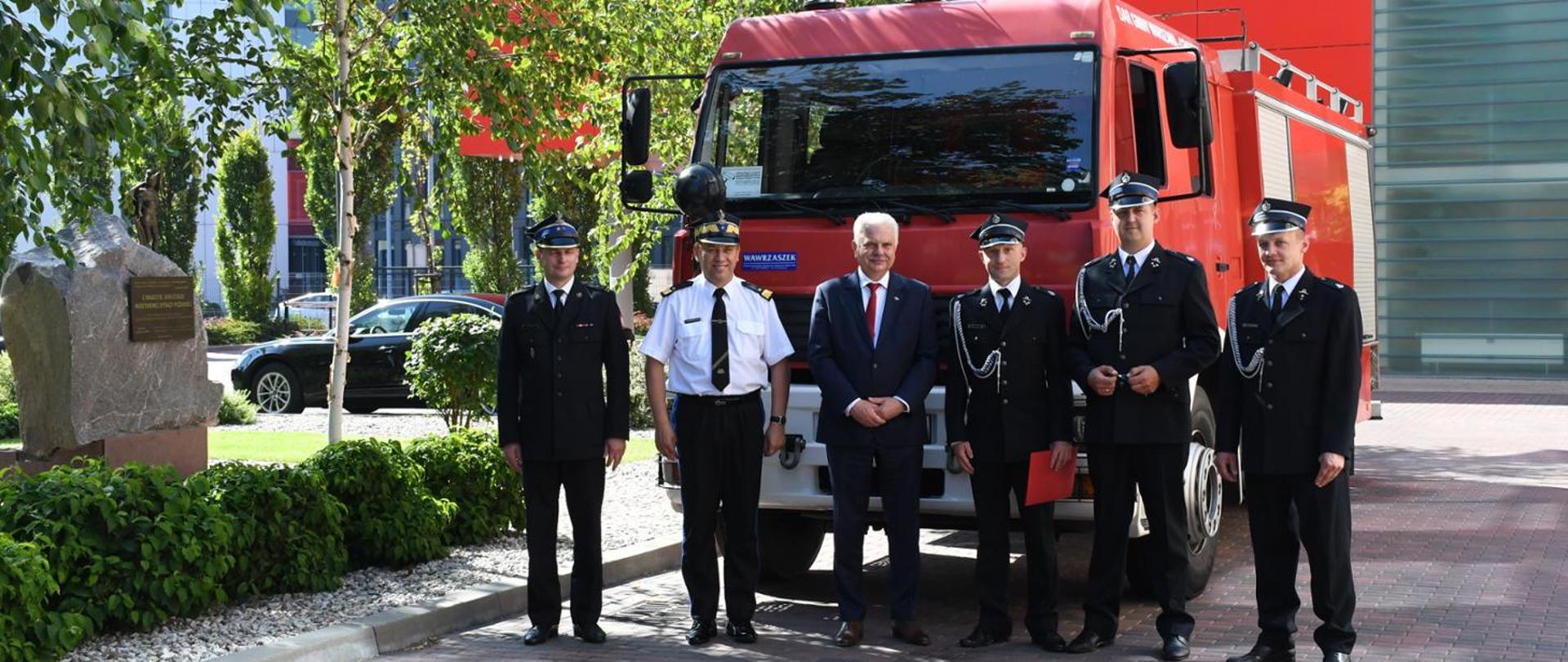 Przekazanie ciężkiego samochodu ratowniczo-gaśniczego MAN jednostce Ochotniczej Straży Pożarnej w Dybowie - strazacy w mundurach galowych w tle samochód pożarniczy