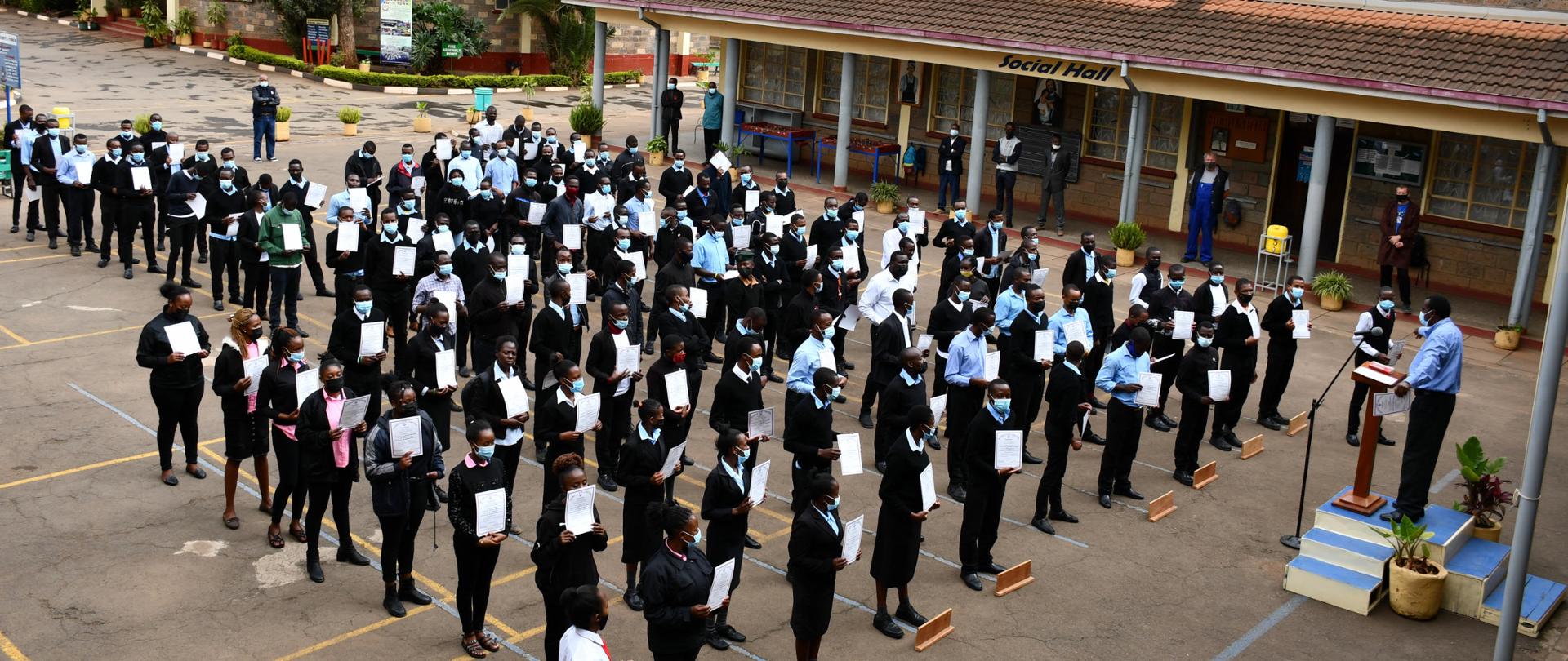 Na placu przed budynkiem szkoły stoją w szeregu chłopcy i dziewczęta ubrani w czerń i biel. W rękach trzymają dyplomy. Przed nimi na błękitnym podwyższeniu przy mikrofonie przemawia mężczyzna w błękitnej koszuli. 