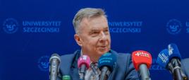 Minister Wieczorek siedzi za stołem na którym stoi kilka mikrofonów, w tle niebieska ścianka z napisem Uniwersytet Szczeciński.
