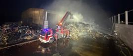 Zdjęcie nocne przedstawiające akcję gaśniczą na składowisku odpadów. Czerwony wóz strażacki z wysuniętą drabiną i koszem operuje nad dymiącą hałdą śmieci obok hali.