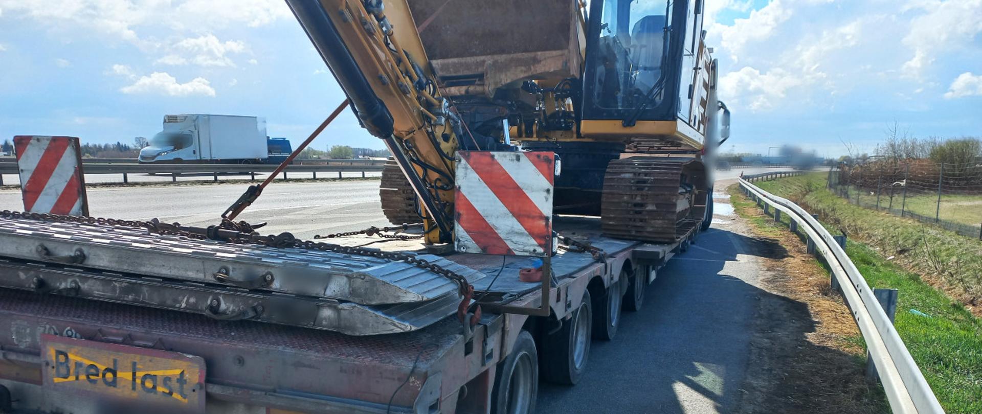 Od lewej: tył naczepy niskopodwoziowej podpiętej do ciągnika siodłowego. Zespołem pojazdów jest przewożona duża koparka gąsienicowa. Zestaw stoi w zatoce kontrolnej przy drodze ekspresowej S8.