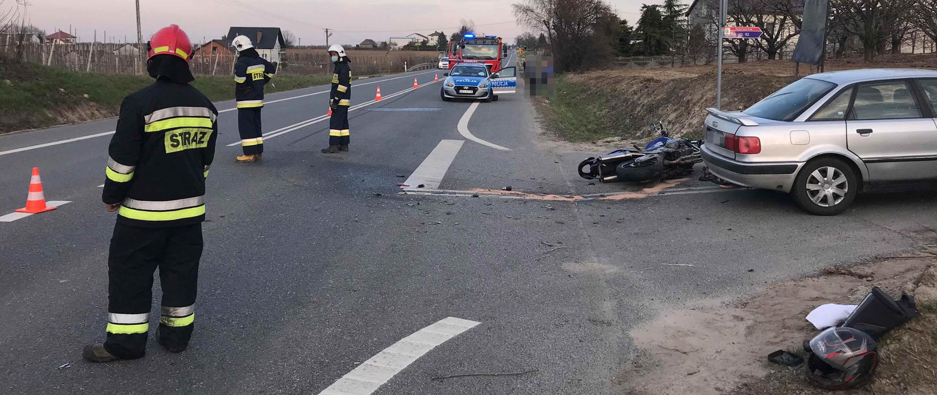 Zdjęcie zrobione na tle drogi krajowej. Na zdjęciu widać jeden samochód osobowy zaparkowany w bocznej drodze i jeden motocykl przewrócony na bok. W oddali widać samochód strażacki i radiowóz Policji . Przy samochodzie i motocyklu , widać rozbite elementy pojazdów i plamę płynów eksploatacyjnych. Pasy jezdni oddzielone są pachołkami ostrzegawczymi przy których stoją strażacy.