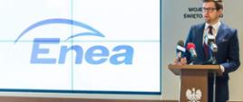Minister Andrzej Śliwka, za jego plecami na ścianie wisi godło. Obok na ekranie wyświetla się logo Enei