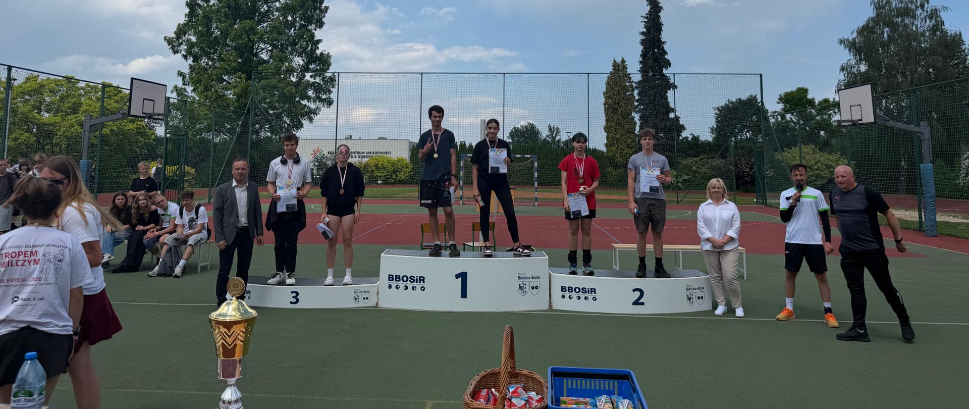 
„Na boisku sportowym stoi podium z miejscami 1, 2 i 3 oraz napisem BBOSiR Bielsko‑Biała. Na podium stoi kilka osób z medalami i dyplomami. Obok stoją organizatorzy i uczestnicy. Na pierwszym planie znajduje się stół z dużym złotym pucharem, dokumentami, sokami i koszem ze słodyczami. W tle widać ogrodzenie boiska, kosze do koszykówki, drzewa i częściowo zachmurzone niebo.”