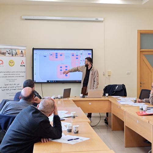 Spotkanie prowadzi stojący mężczyzna, który pokazuje schemat na prezentacji. Obok niego baner z logo Polska pomoc i PCPM. Przy długicj stołach siedzi 5 osoób, które słucha prezentacji i robi notatki. 