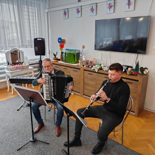 Na zdjęciu widać dwóch mężczyzn. Obaj siedzą na jasnych krzesłach. Przed nimi stoją pulpity na nuty. Mężczyzna siedzący z prawej strony jest ubrany na czarno i gra na czarnym klarnecie. Mężczyzna siedzący z lewej strony jest ubrany w niebieskie spodnie, białą koszulę, ciemną marynarkę i gra na czarnym akordeonie. W tle widać brązową komodę z dekoracją świąteczną, telewizor zawieszony na jasnej ścianie, czarny instrument klawiszowy na statywie oraz kolumnę głośnika na statywie.