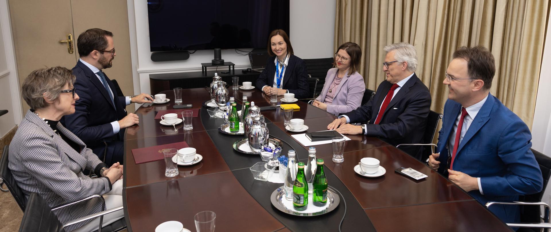 Six people take part in a meeting around a conference table set with cups, glasses, and bottles of water. The participants, dressed in formal business attire, are engaged in discussion in a small conference room.
