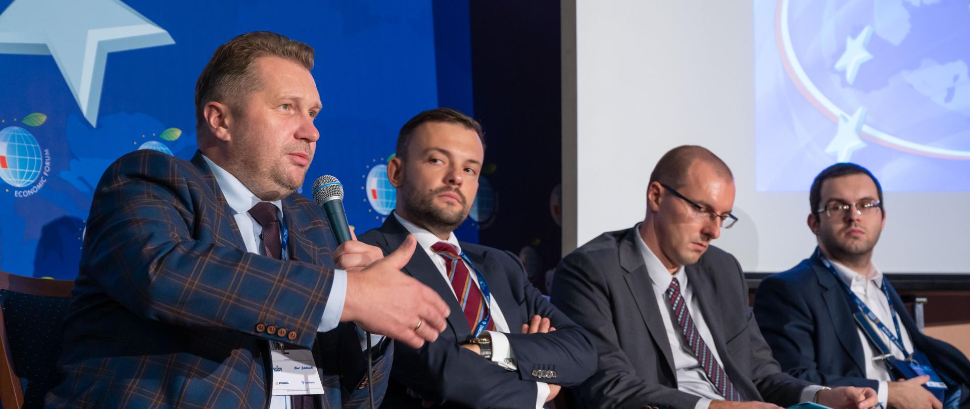 Na zdjęciu minister Czarnek i kilka innych osób siedzą na krzesłach na tle niebieskiej ściany, minister trzyma mikrofon i mówi do niego.