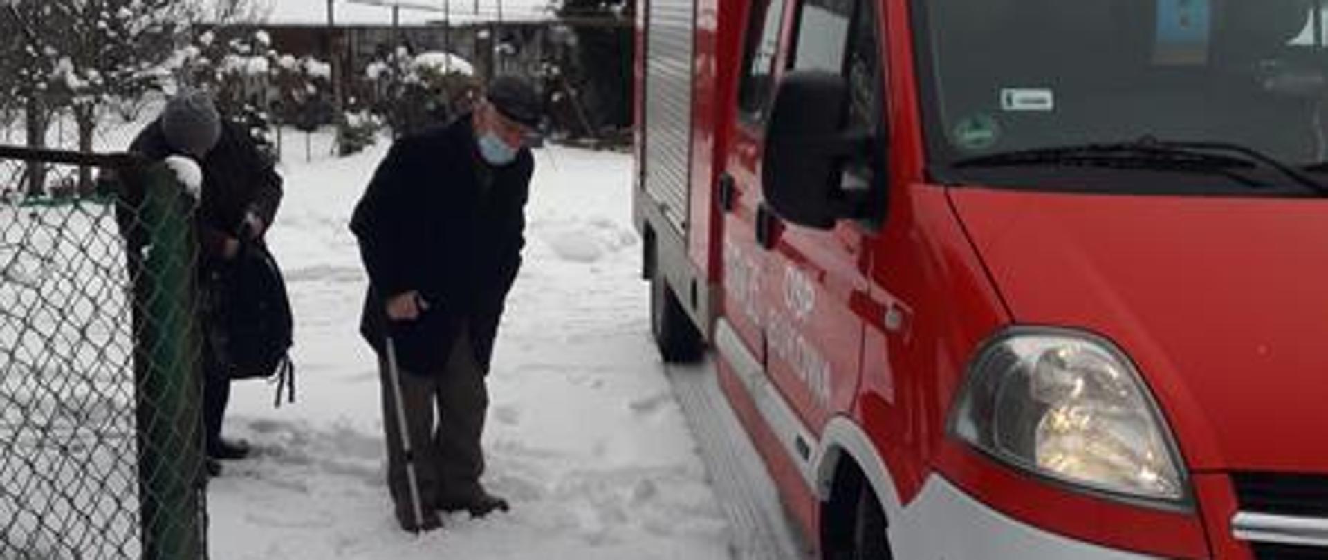 Na posesji znajduje się samochód pożarniczy koloru czerwonego. Dwie starsze osoby znajdują się obok pojazdu przygotowując się do transportu samochodem do punktu szczepień.