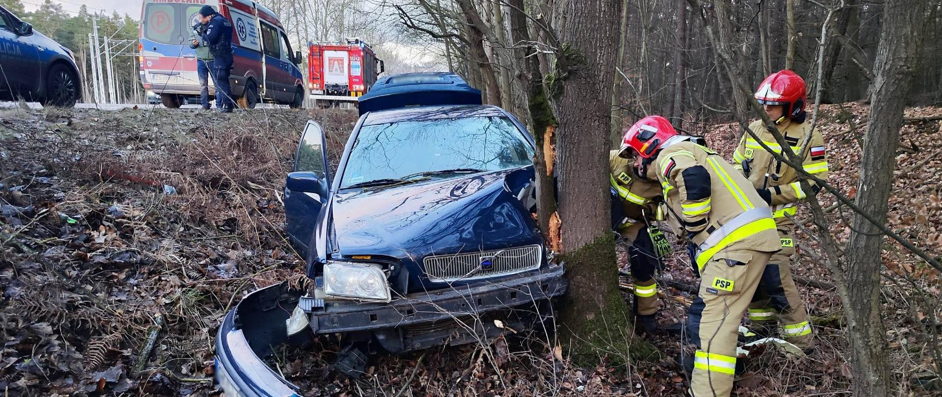 Zdjęcie przedstawia uszkodzony samochód marki Volvo w wyniku uderzenia w drzewo. Samochód stoi na ziemi pokrytej liśćmi, przy drzewie. Po prawej stronie widać 3 strażaków w trakcie działań. Po lewej stronie w tle widać karetkę pogotowia wraz z samochodem pożarniczym oraz funkcjonariusza Policji podczas czynności.